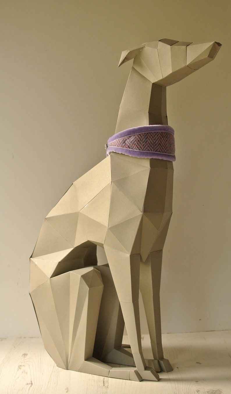 May include: A beige papercraft sculpture of a greyhound dog sitting with its legs crossed. The dog is wearing a purple and pink patterned collar.