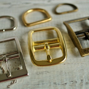 May include: Five different metal buckles in various sizes and finishes. The buckles are made of silver, gold, and brass. They are all rectangular in shape and have a simple design.