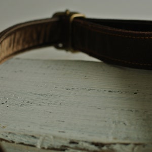 May include: A brown leather belt with a brass buckle. The belt is lying on a white wooden surface.