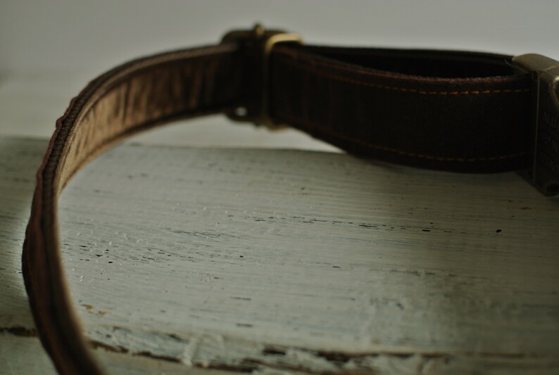 May include: A brown leather belt with a brass buckle. The belt is lying on a white wooden surface.