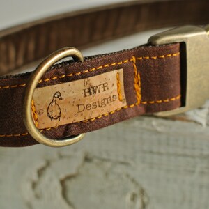 May include: Brown canvas dog collar with a cork tag that reads "HWR Designs" and a duck illustration. The collar has a brass buckle and D-ring.