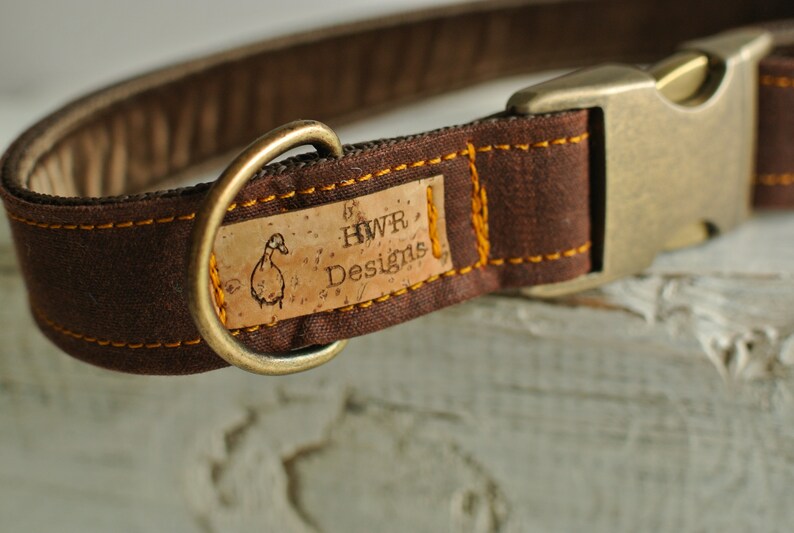 May include: Brown canvas dog collar with a cork tag that reads "HWR Designs" and a duck illustration. The collar has a brass buckle and D-ring.