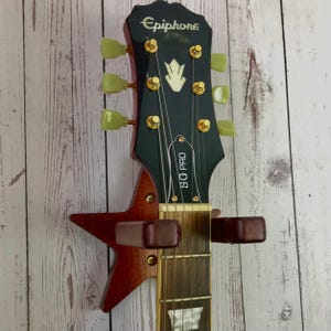Op de afbeelding: Een rode Epiphone SG Pro elektrische gitaar met gouden hardware en een stervormige wandbevestiging. De gitaar heeft een zwarte kop met het Epiphone-logo en de woorden "SG Pro" gedrukt op de voorkant.