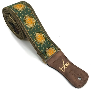 May include: A guitar strap with a green and gold sun design. The strap has a brown leather end piece with gold text. The strap is made of a woven material and has metal hardware.