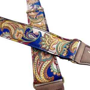 May include: A patterned guitar strap with a paisley design. The strap displays a vibrant pattern in blue, yellow, red, and green. The ends are made of brown leather and have metal hardware.