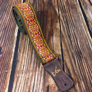 Könnte beinhalten: Ein grüner, orangefarbener und violetter Gitarrengurt mit einem braunen Lederende. Das Lederende hat das Wort "Van" geprägt.