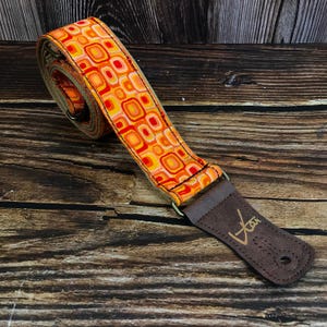 May include: A guitar strap with a retro design. The strap has a repeating pattern of orange, yellow, and red squares and circles. The end piece is brown leather with a gold logo.