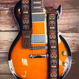 Op de afbeelding: Een bruine leren gitaarband met een kleurrijk tribal patroon met gestileerde gezichten. De band is bevestigd aan een sunburst elektrische gitaar met chromen hardware.