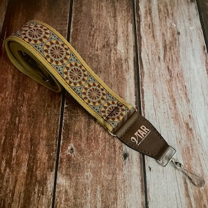 May include: A brown and yellow guitar strap with a geometric pattern and a brown leather end with the text "2TAR" printed on it.