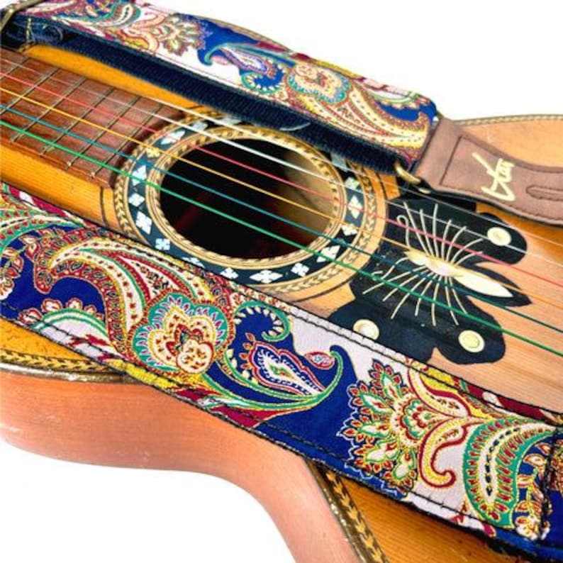 May include: An acoustic guitar with a vibrant paisley guitar strap. The strap displays a blue base with intricate red, yellow, and green paisley patterns. The guitar's wooden body and soundhole are visible, with strings and a decorative rosette.