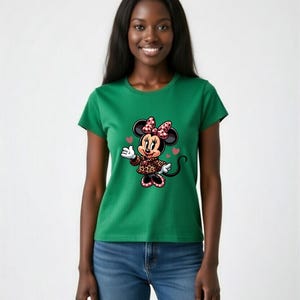 May include: Green t-shirt featuring a Minnie Mouse graphic. The cartoon character is wearing a leopard print jacket and a bow with pink polka dots. The design includes hearts. The model is wearing blue jeans.