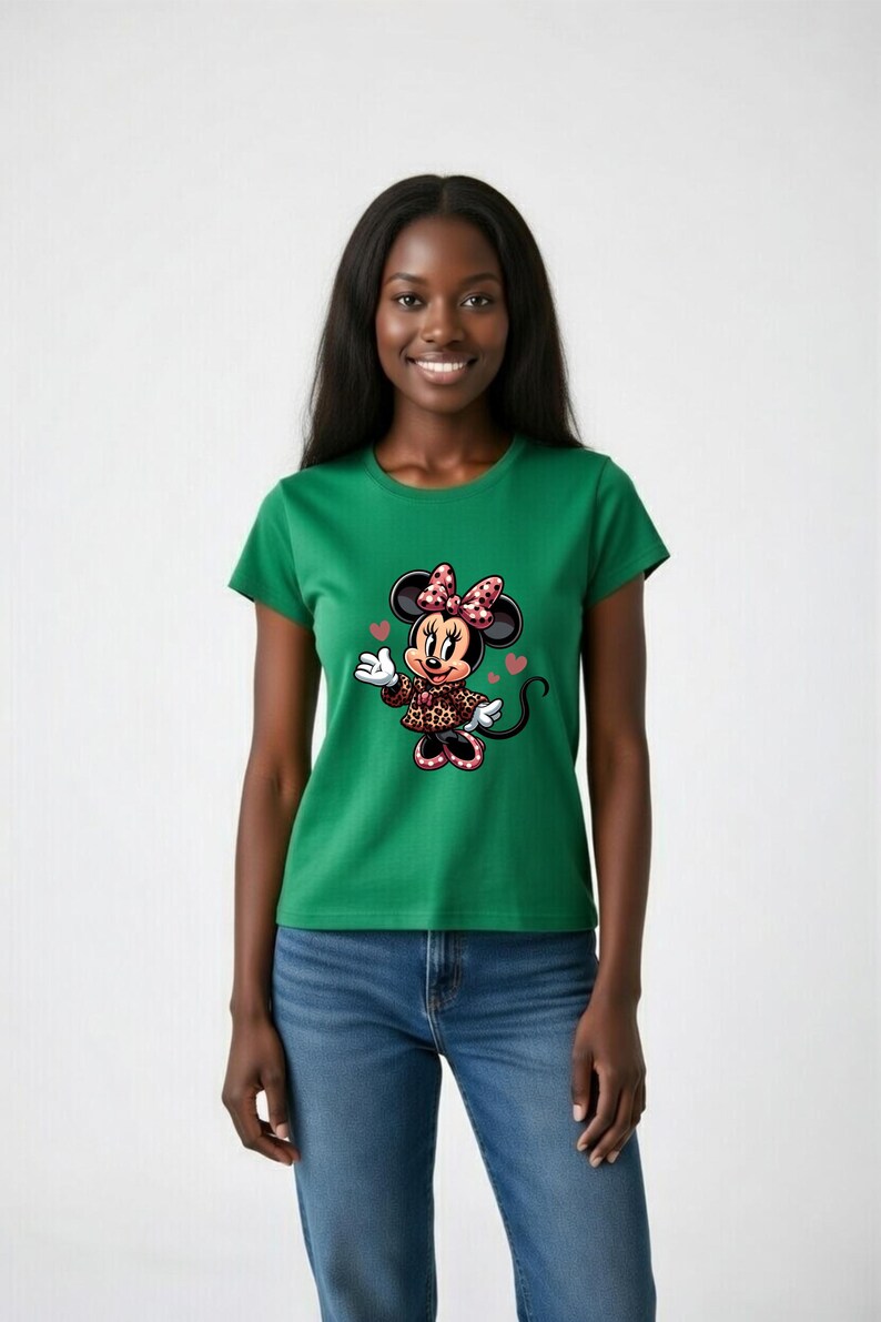May include: Green t-shirt featuring a Minnie Mouse graphic. The cartoon character is wearing a leopard print jacket and a bow with pink polka dots. The design includes hearts. The model is wearing blue jeans.