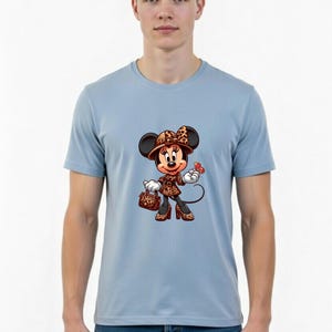May include: Light blue t-shirt featuring a cartoon Minnie Mouse character in a stylish outfit. Minnie is wearing a leopard print hat, coat, and shoes, carrying a matching purse, and holding a red butterfly. The shirt is worn by a person.