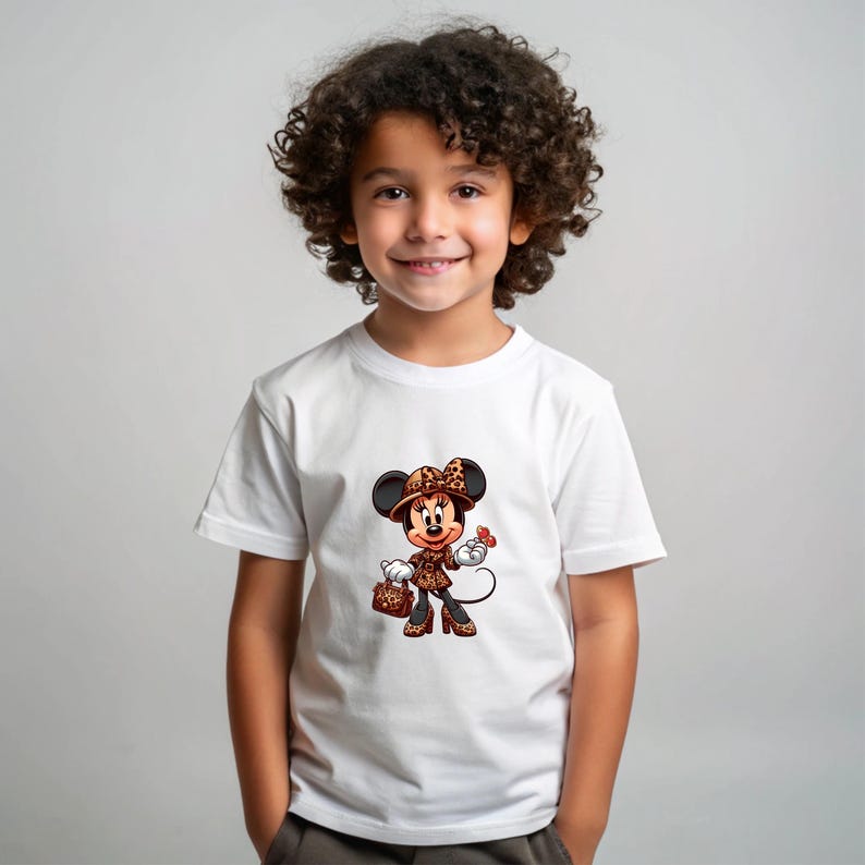 May include: A white t-shirt featuring a cartoon Minnie Mouse character in a leopard print outfit, including a hat, jacket, and purse. The character is holding a red flower. The child is smiling.