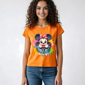May include: Orange t-shirt featuring a cartoon Minnie Mouse design. The graphic shows Minnie peeking out of a zippered pocket with colorful paint splatter accents. The shirt is paired with blue jeans.
