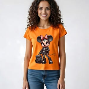Peut inclure: T-shirt orange avec un personnage de dessin anim&eacute; Minnie Mouse en tenue noire, tenant un sac &agrave; dos. Le personnage a un n&oelig;ud rouge &agrave; pois blancs. Le mod&egrave;le porte un jean bleu.