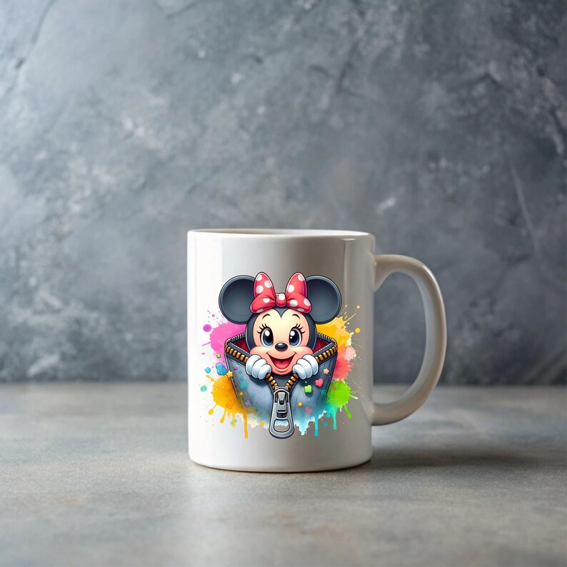 May include: White ceramic mug featuring a cartoon Minnie Mouse design. Minnie Mouse is wearing a red bow and is inside a zippered jacket with colorful paint splatter accents. The mug has a curved handle.