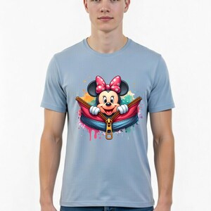 May include: Light blue t-shirt featuring a colorful graphic of Minnie Mouse emerging from a zippered pouch. Minnie Mouse is wearing a pink polka-dot bow. The design includes colorful paint splatters.