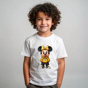May include: White t-shirt featuring a cartoon Minnie Mouse character wearing a yellow hoodie, black shorts, and sneakers. The character has a yellow bow and black ears. The shirt is worn by a young child.