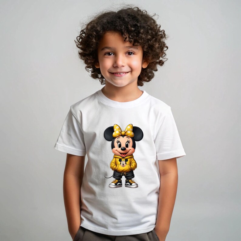 May include: White t-shirt featuring a cartoon Minnie Mouse character wearing a yellow hoodie, black shorts, and sneakers. The character has a yellow bow and black ears. The shirt is worn by a young child.