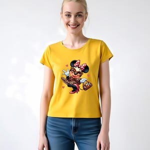 May include: Mustard yellow t-shirt featuring a cartoon Minnie Mouse in a leopard print outfit, red bow, and sunglasses. The graphic includes hearts and a purse. The model is wearing blue jeans.