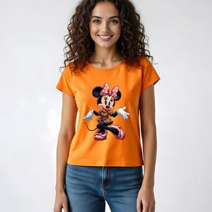 May include: Orange t-shirt featuring a Minnie Mouse graphic. The cartoon character is depicted in a playful pose, wearing a polka-dot bow, a leopard print top, and pink shoes. The model is wearing blue jeans.