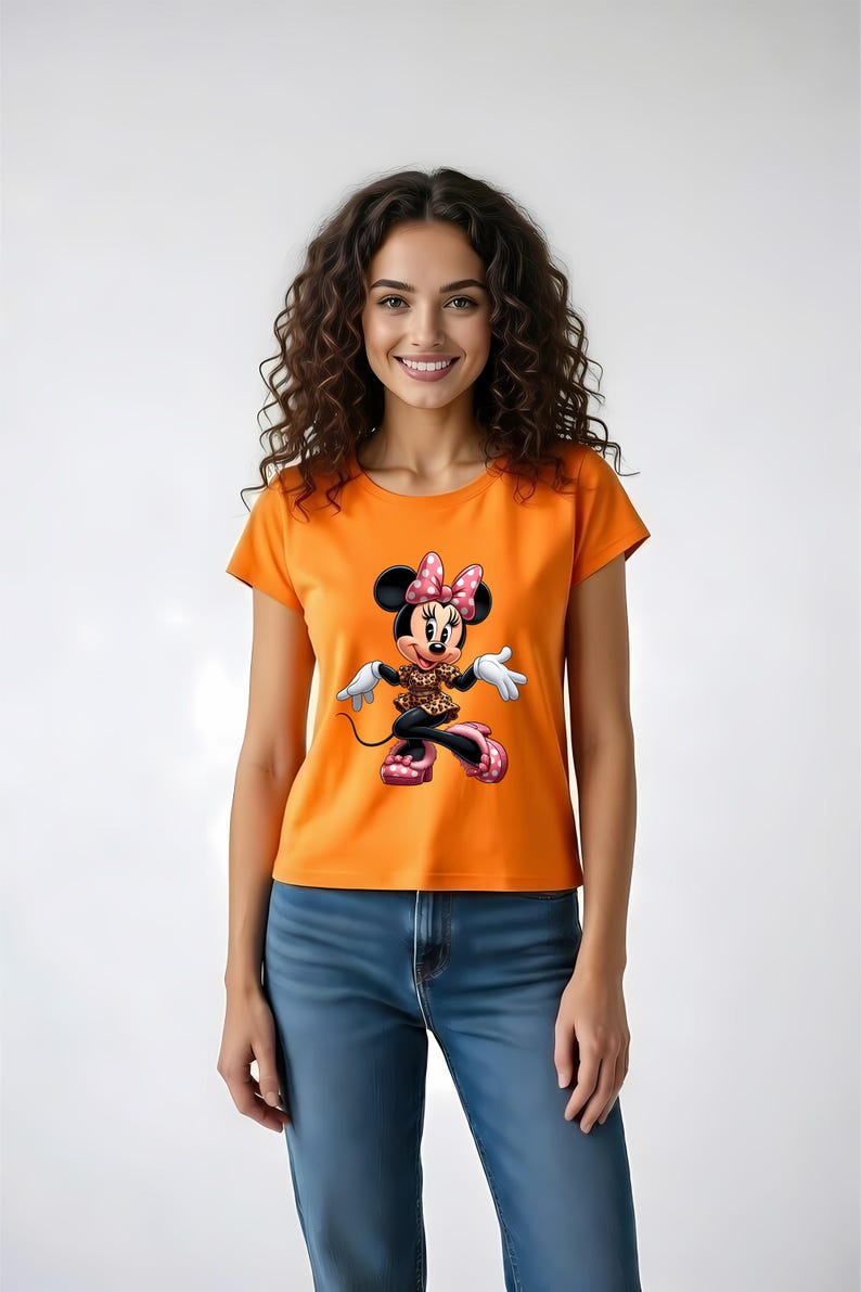 May include: Orange t-shirt featuring a Minnie Mouse graphic. The cartoon character is depicted in a playful pose, wearing a polka-dot bow, a leopard print top, and pink shoes. The model is wearing blue jeans.