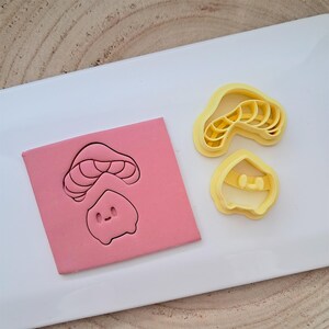Pode incluir: Dois cortadores de biscoitos de pl&aacute;stico amarelo, um com a forma de um cogumelo e o outro com a forma de um cogumelo com rosto. O cortador de cogumelo &eacute; em camadas para criar um design detalhado. Os cortadores est&atilde;o sobre uma superf&iacute;cie branca.