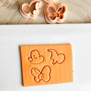 May include: Pink plastic cookie cutters in the shapes of a bow, a mouse head, and a character's head. The shapes are pressed into an orange block of clay. The cutters are on a wooden surface.