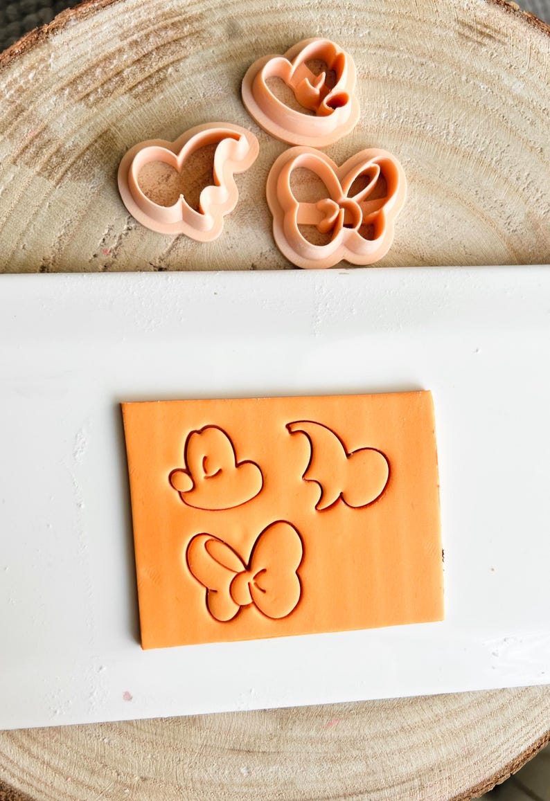 May include: Pink plastic cookie cutters in the shapes of a bow, a mouse head, and a character's head. The shapes are pressed into an orange block of clay. The cutters are on a wooden surface.