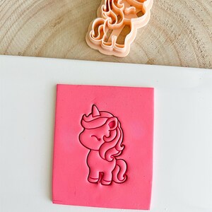 May include: A light orange unicorn cookie cutter next to a pink block with a unicorn imprint. The unicorn design features a horn, flowing mane, and a smiling face. The cookie cutter is made of plastic and is designed for baking.