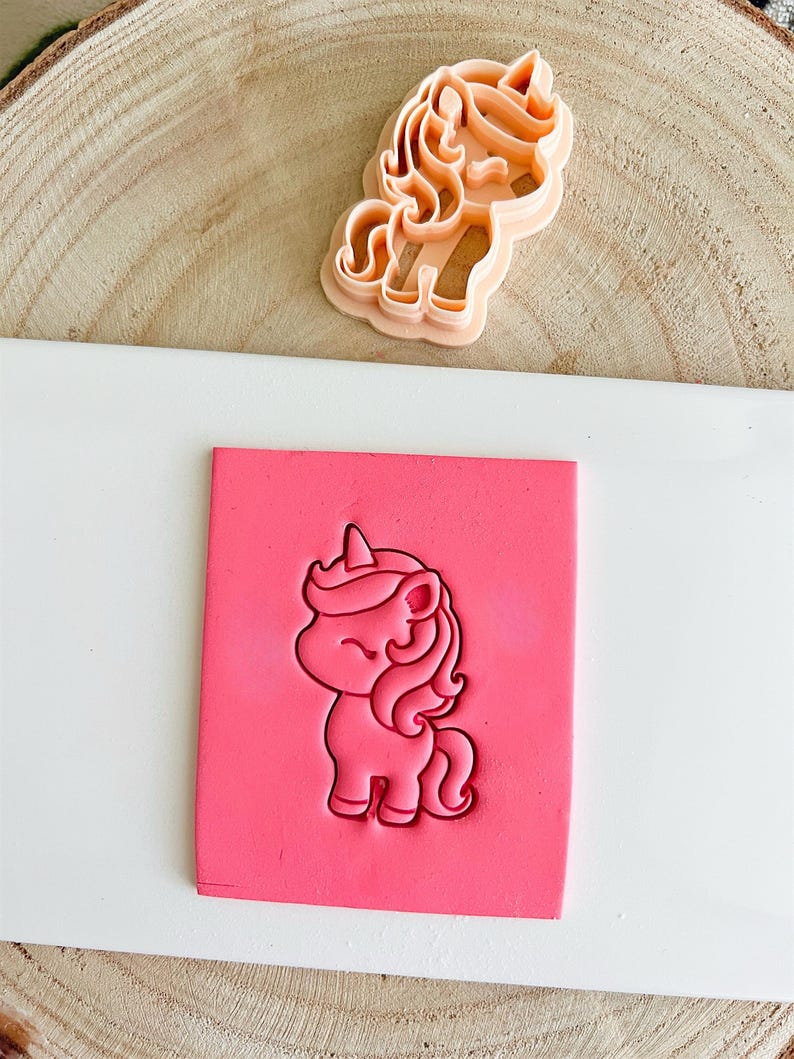 May include: A light orange unicorn cookie cutter next to a pink block with a unicorn imprint. The unicorn design features a horn, flowing mane, and a smiling face. The cookie cutter is made of plastic and is designed for baking.