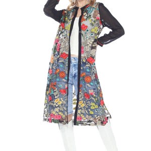 May include: A long, sheer floral embroidered duster coat with black sleeves and trim. The coat features a colorful flower pattern and is worn over a white top and denim shorts. White boots complete the outfit.