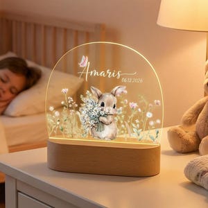 May include: A decorative night light featuring a bunny holding flowers, with the name "Amaris" and the date "06.12.2026" etched on the clear acrylic. The light sits on a wooden base and is placed on a white nightstand in a child's bedroom.