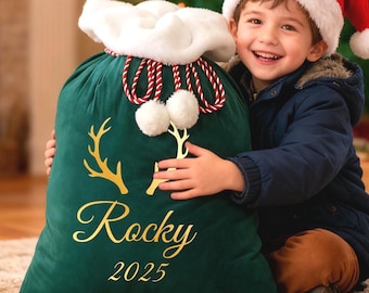 May include: A dark green velvet Christmas gift bag with gold antlers and the name "Rocky" and the year "2025" printed on it. The bag has a white fluffy trim and red and white rope ties with white pom-poms. A child wearing a Santa hat is holding the bag.