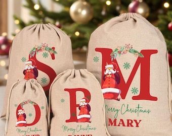 May include: Four beige Christmas gift bags with red letters and Santa Claus illustrations. The bags are personalised with names and the words "Merry Christmas". The bags are different sizes and have a drawstring closure.