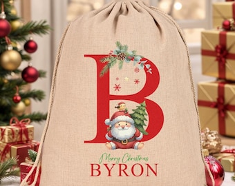 May include: A beige Christmas gift bag with a red letter "B" design, a Santa Claus graphic, and the text "Merry Christmas BYRON". The bag is made of a drawstring material and is surrounded by festive decorations.