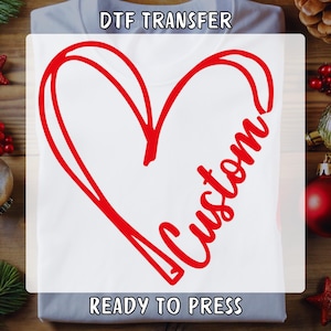 May include: A white t-shirt with a red heart design and the word "Custom" written inside the heart. The text "DTF Transfer" is at the top of the shirt and "Ready to Press" is at the bottom.