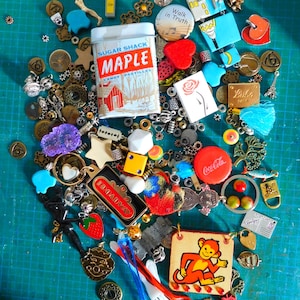 Può includere: Una collezione di ciondoli e charms colorati, tra cui una piccola scatola di latta con le parole "Sugar Shack Maple Candy Pastilles", un tappo di bottiglia Coca-Cola, un piccolo charm di scimmia e una varietà di altri charms e perline.