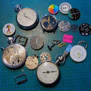 May include: A collection of vintage watch parts and mechanisms, including a Mickey Mouse watch face, a stopwatch, a silver watch with a black face, and a variety of gears and watch hands.