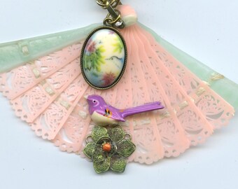 May include: A pink and green fan-shaped pendant with a floral design, a purple bird, and a green flower with a red center.