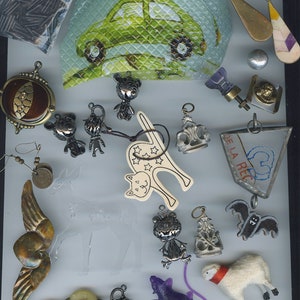 Può includere: Assortimento di ciondoli e pendenti, tra cui un gatto, un cervo, una pecora, un pipistrello e un topo viola. Ci sono anche diversi ciondoli dall'aspetto vintage, tra cui una piccola auto, una corona e un gatto con le stelle. I ciondoli sono esposti su una superficie grigio chiaro.
