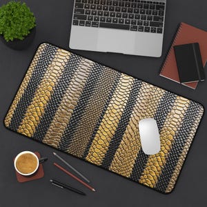 May include: A rectangular desk mouse pad with a black border. The pad features a pattern of alternating gold and black snake-skin-like stripes. A white computer mouse sits on the pad, with a laptop, notebook, and coffee cup in the background.