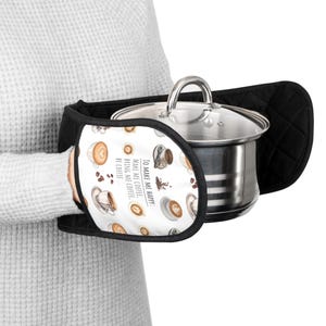 Puede incluir: Manoplas negras con una sección estampada en blanco con tazas de café y el texto "TO MAKE ME HAPPY: MAKE ME COFFEE. BRING ME COFFEE. BE COFFEE." Una olla de acero inoxidable con tapa de cristal es sostenida por las manoplas.