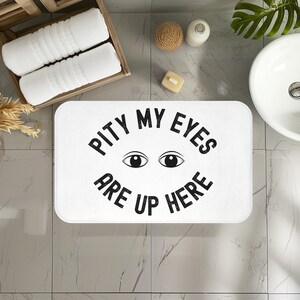 May include: White rectangular bath mat with the text "PITY MY EYES ARE UP HERE" in black. The mat features two black eye illustrations. The mat is in a bathroom setting with white towels and a sink.