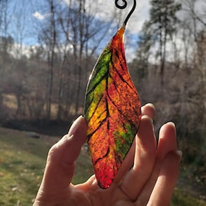 May include: A colorful, leaf-shaped ornament with a black wire hook. The leaf features vibrant hues of red, orange, yellow, and green, resembling autumn foliage. The ornament is held up against a backdrop of trees and a bright sky.