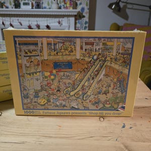 May include: A 1000 piece jigsaw puzzle box featuring a colourful illustration of a busy shopping mall with escalators, shoppers, and a sign that reads "Welcome to the Mall, Shop till you drop!"