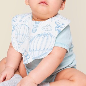 May include: A baby wearing a light blue onesie with a white bib featuring a blue hot air balloon pattern. The bib has a scalloped edge and a hook and loop closure.