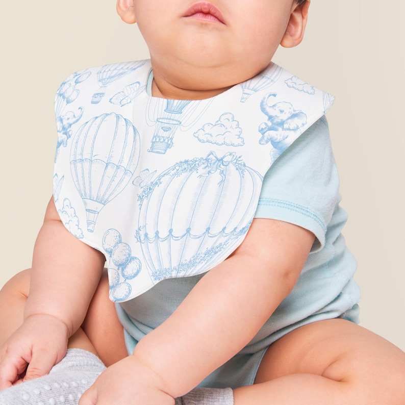 May include: A baby wearing a light blue onesie with a white bib featuring a blue hot air balloon pattern. The bib has a scalloped edge and a hook and loop closure.
