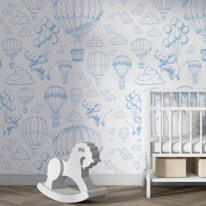 May include: A white nursery wall with a blue and white wallpaper featuring hot air balloons, clouds, butterflies, and elephants. A white rocking horse and a white crib are in the foreground.